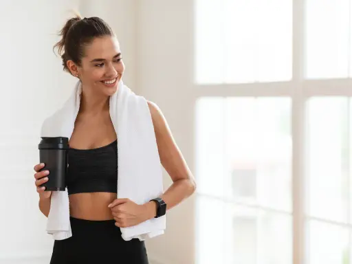 Lächelnde Frau mit Handtuch und Shaker nach dem Training. Sie fragt sich, wie viel Kreatin pro Tag für optimale Ergebnisse.