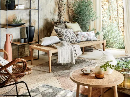 Un salon sans canapé, avec un banc tressé, des coussins décoratifs, un fauteuil en rotin et une étagère murale.