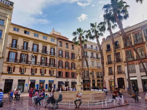 Sonniger Platz in der **Malaga Altstadt** mit einem Brunnen, Palmen und bunten Gebäuden. Menschen flanieren und genießen die Atmosphäre.