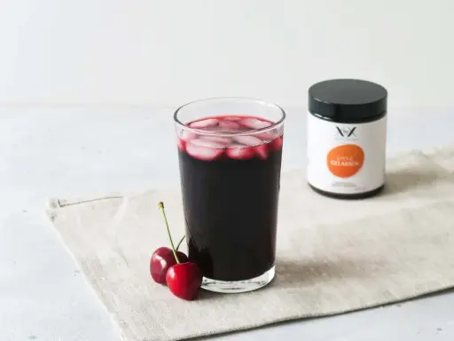 Ein Glas dunkler Sauerkirschsaft mit Eiswürfeln und zwei Kirschen daneben. Eine Dose mit der Aufschrift "Ganz Gelassen" steht im Hintergrund. Die positive sauerkirschsaft wirkung wird hier visuell dargestellt.