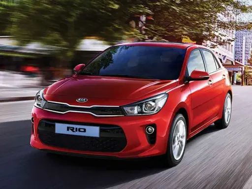 Un hatchback rojo Kia Rio se desplaza por una calle urbana, con edificios y árboles borrosos por el movimiento.