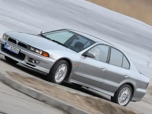 Srebrny Mitsubishi Galant z gracją pokonuje zakręt. Kierowca w okularach pewnie prowadzi auto.