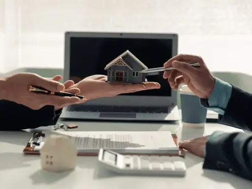 Deux mains présentent une maquette de maison, symbolisant comment devenir investisseur immobilier. Un contrat et un ordinateur sont visibles.