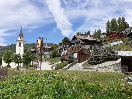 A picturesque village in Chamois, Italy, with a church tower, wooden chalets, and lush greenery. Scopri cosa vedere in questo incantevole borgo alpino.