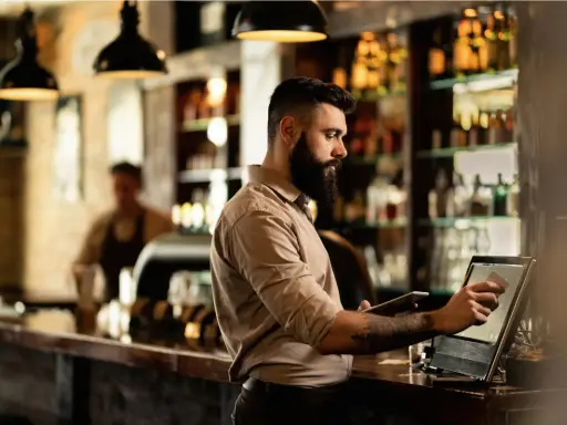 Hombre con barba gestionando un bar, usando una tablet y una pantalla táctil para procesar pagos.