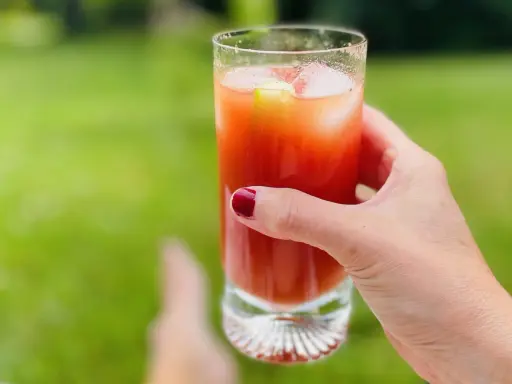 A hand with red nail polish holds a tall glass of Bloody Mary with ice and a cucumber slice, outdoors.