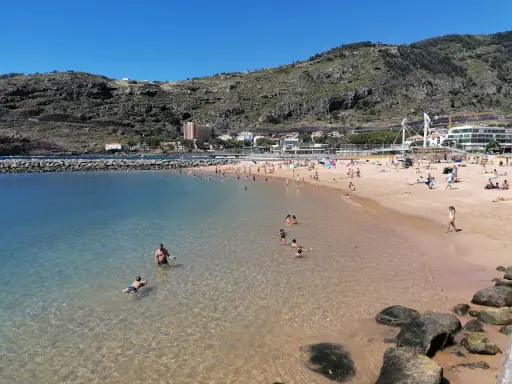 Sonniger Strand in Machico, Madeira, mit vielen Menschen, die im klaren Wasser planschen und sich am Sandstrand sonnen.