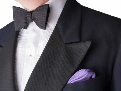 A man wears a black tuxedo with a pleated white shirt and a black bow tie. A purple pocket square adds a pop of color. This outfit answers the question: can you wear a tie with a tux? (No, a bow tie is traditional.)