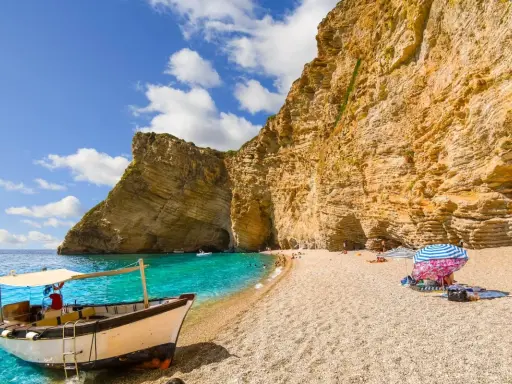 Ein Boot liegt am Korfu Strand vor einer beeindruckenden Felswand. Türkises Wasser und ein sonniger Himmel laden zum Verweilen ein.