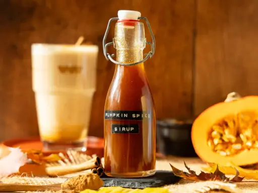 Flasche mit goldbraunem Pumpkin Spice Sirup, daneben ein Latte Macchiato, Kürbishälften und Herbstlaub.