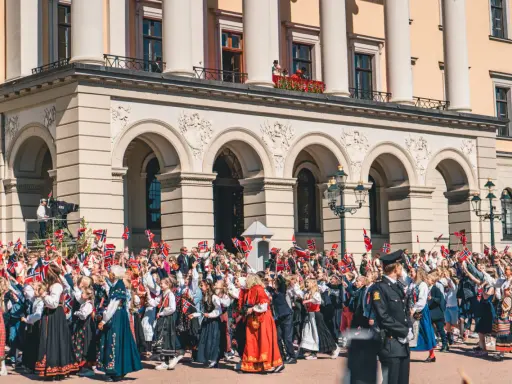 Tłum ludzi, w tym dzieci, świętuje, machając norweskimi flagami. Wiele osób nosi tradycyjny strój norweski, ozdobiony haftami i kolorowymi wzorami.