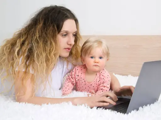 Kobieta z dzieckiem na rękach pracuje na laptopie, zastanawiając się, czy na urlopie wychowawczym można pracować.