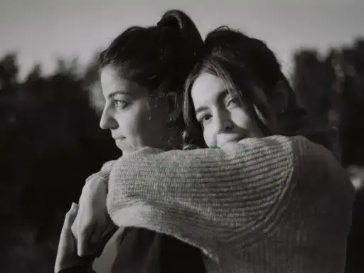 Dos mujeres se abrazan, una mirando al horizonte con **miedo al abandono**, la otra la abraza con ternura.