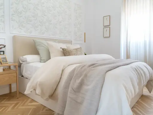 Dormitorios con papel pintado de hojas verdes en el cabecero. Cama con ropa de cama blanca y manta gris. Mesita de noche de madera.