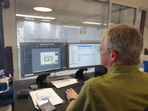 Mann am Computer, der Baupläne nach DIN 4426 erstellt. Auf dem Bildschirm sind CAD-Zeichnungen und eine Liste von Bauteilen zu sehen.