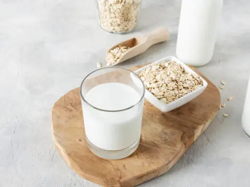 Ein Glas cremiger Hafermilch steht auf einem Holzbrett, daneben Schüsseln mit Haferflocken. Hafermilch gesund und lecker!