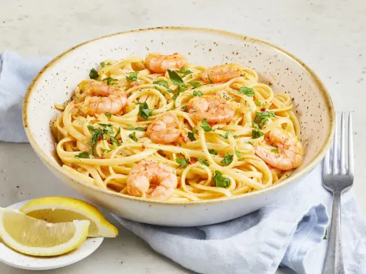 Eine Schüssel mit Spaghetti mit Garnelen, garniert mit Petersilie und Chiliflocken. Dazu Zitronenspalten und eine Gabel.