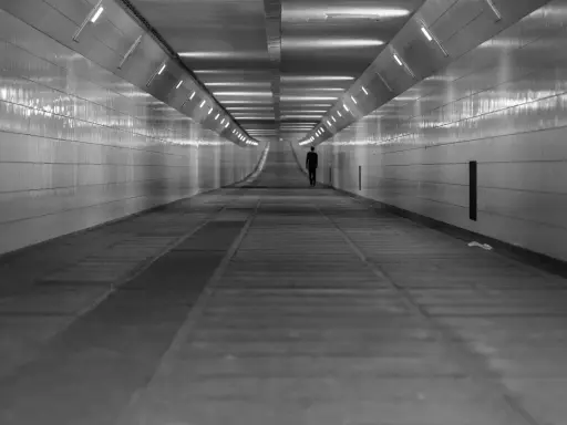 Człowiek w garniturze idzie przez długi, pusty tunel. Światła na ścianach tworzą wrażenie nieskończoności, jakby wchodził w szarą strefę.