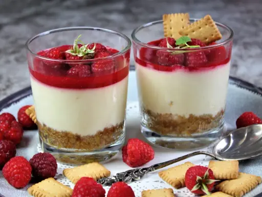 Zwei Gläser mit kalorienarmen Desserts im Glas: cremige Schicht, Himbeersoße und Keksboden. Garniert mit frischen Himbeeren und Keksen.