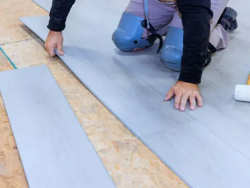 Man con rodilleras coloca cuidadosamente tablones de suelo laminado gris sobre una base de madera.
