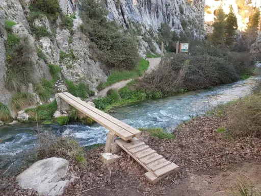 Puente de madera sobre un río caudaloso, parte de la ruta del zarzalar. Senderos y vegetación agreste flanquean el camino.