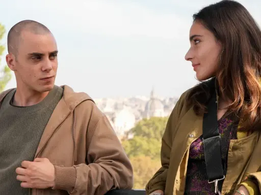 Due giovani si guardano intensamente, come se fossero in una "casa degli sguardi". Lui, rasato, lei con lunghi capelli castani.