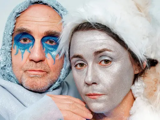 Dwoje ludzi z artystycznym makijażem. Mężczyzna w niebieskim kapturze z błyszczącymi detalami, kobieta w białym futrzanym nakryciu głowy. Ich twarze pokryte są srebrnym i niebieskim brokatem, niczym serce ze szkła.