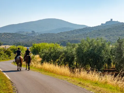 Due cavalieri godono di una **passeggiata a cavallo toscana** tra ulivi e colline, con un castello in lontananza.
