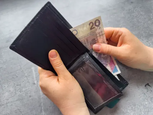 Ręce wkładają banknot 20 zł do portfela. Dowiedz się, komu przysługuje zasiłek dla bezrobotnych.
