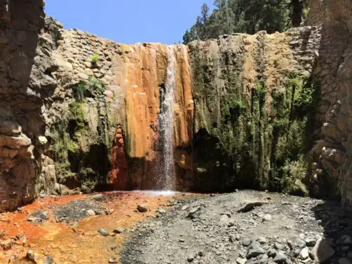 La cascada de colores la Palma, con sus tonos ocres y verdes, cae sobre rocas y vegetación exuberante en un cañón.