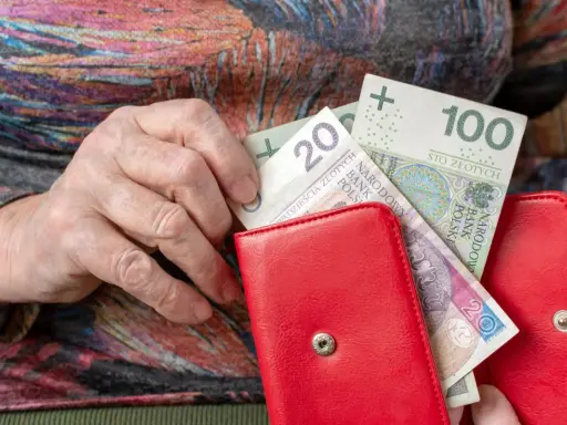 Dłoń starszej osoby wkłada pieniądze do czerwonego portfela. Może to być dodatek mieszkaniowy.