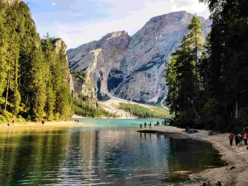 Sentiero da Valdaora al Lago di Braies: acque turchesi, montagne imponenti e pinete rigogliose. Turisti passeggiano sulla riva sabbiosa.