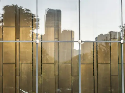 Reflejos dorados de árboles y edificios en un **cristal blindado** moderno, con estructura metálica visible.
