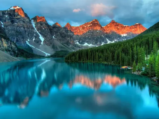 Turkusowe jezioro odbija ośnieżone szczyty gór o zachodzie słońca. W tle gęsty las iglasty. Park narodowy Banff zachwyca.