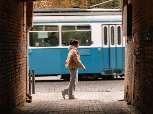 Ragazza con piumino beige cammina su un vicolo di mattoni, mentre un tram blu sfreccia sullo sfondo. Una perfetta foto aesthetic da fare.