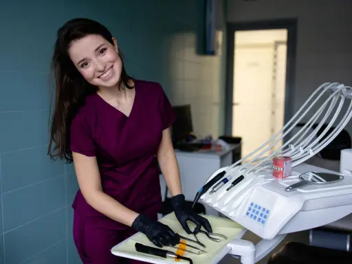 Uśmiechnięta asystentka stomatologiczna w bordowym uniformie przygotowuje narzędzia na tacce obok fotela dentystycznego.