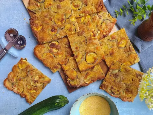 Fette dorate di schiacciata di zucchine, condita con erbe aromatiche, su un tagliere. Accanto, una zucchina fresca, farina di mais e un rotella taglia pizza.