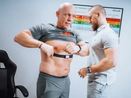 Mann legt Brustgurt für Muskelerhalt Training an, Trainer hilft.