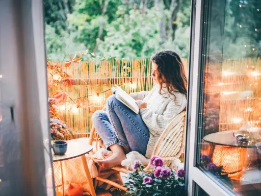 Przytulny jesienny balkon. Dziewczyna w swetrze czyta książkę, otoczona jesiennymi liśćmi i ciepłym światłem lampek.