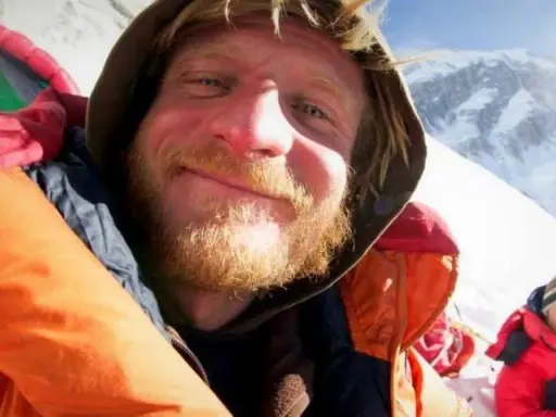 Uśmiechnięty alpinista w ciepłej kurtce, z brodą, na tle ośnieżonych gór. Może to być jego lek na chorobę wysokościową.