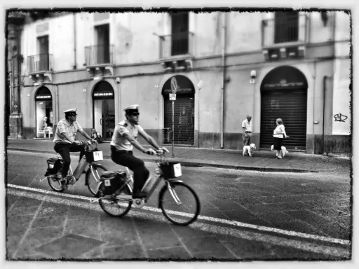 Due agenti in bicicletta pattugliano una strada italiana. Sul marciapiede, una coppia passeggia con i loro cani. Street photography Italia.