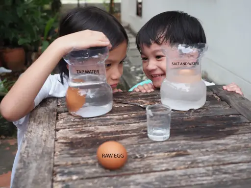 Niños observan un huevo flotando en agua salada y otro hundido en agua normal, parte de sus experimentos con agua.