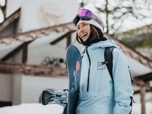 Uśmiechnięta snowboardzistka w jasnoniebieskim anoraku i goglach Anon. Gotowa na stok, trzyma deskę Burton.