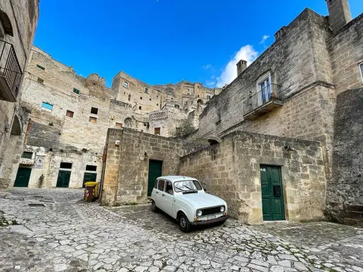 Un'auto d'epoca bianca parcheggiata tra le antiche case scavate nella roccia dei Sassi di Matera, sotto un cielo azzurro.