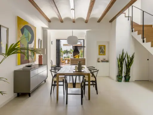 Comedor moderno en una casa entre medianeras, con vigas de madera en el techo, mesa de madera, sillas negras y una escalera de madera.