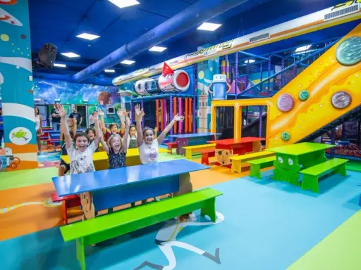 Un grupo de niños alegres levanta las manos en un vibrante centro comercial con juegos para niños, con toboganes, túneles y coloridos muebles.