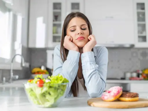 Femme triste face à une salade et des donuts, pensant aux aliments à éviter pour la dermite séborrhéique.