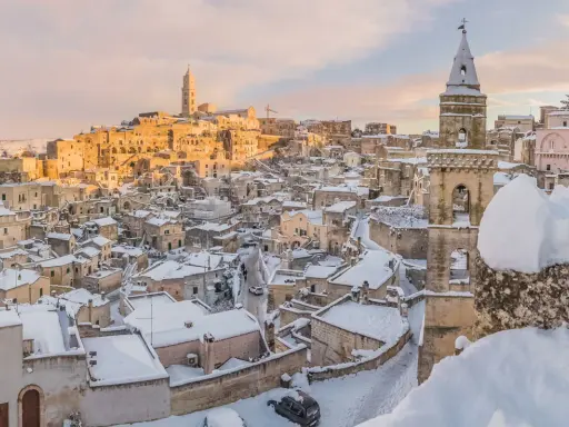 Matera innevato, un incanto tra i **posti da visitare in inverno Italia**. Case antiche e chiese sotto la neve, con il sole che illumina il paesaggio.
