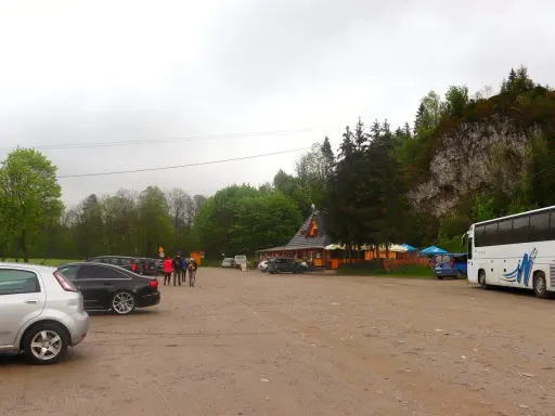 Parking przy Wąwozie Homole. Samochody, autokar i budynek zadaszony, otoczony drzewami, u podnóża skalnego zbocza.