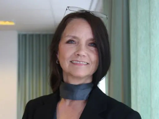 En kvinna med glasögon på huvudet ler. Hon bär en svart kavaj och en grå scarf.